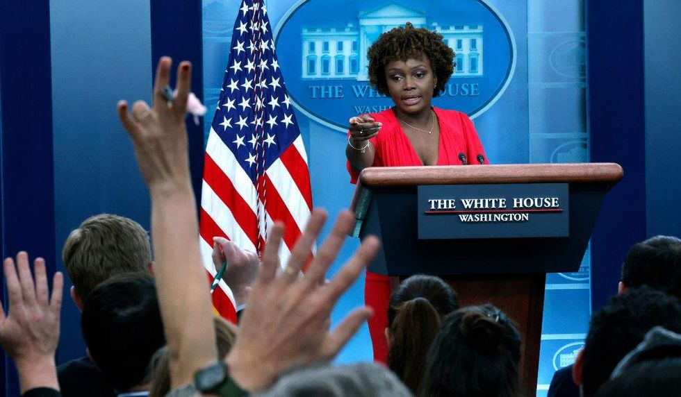 karine jean-pierre at the white house briefing room lectern gesturing to reporters with their hands raised