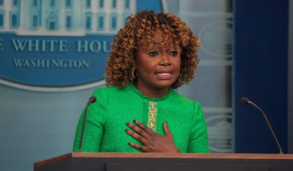 karine jean-pierre wearing a green suit. she's gesturing to her chest as she speaks to the white house press corps