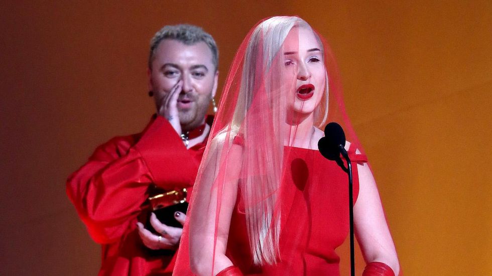 Kim Petras at the microphone accepting her Grammy with Sam Smith behind her supporting her.