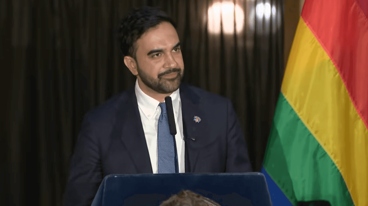 Mayor Zohran Mamdani speaks into a microphone and stands before a rainbow LGBTQ+ Pride flag.