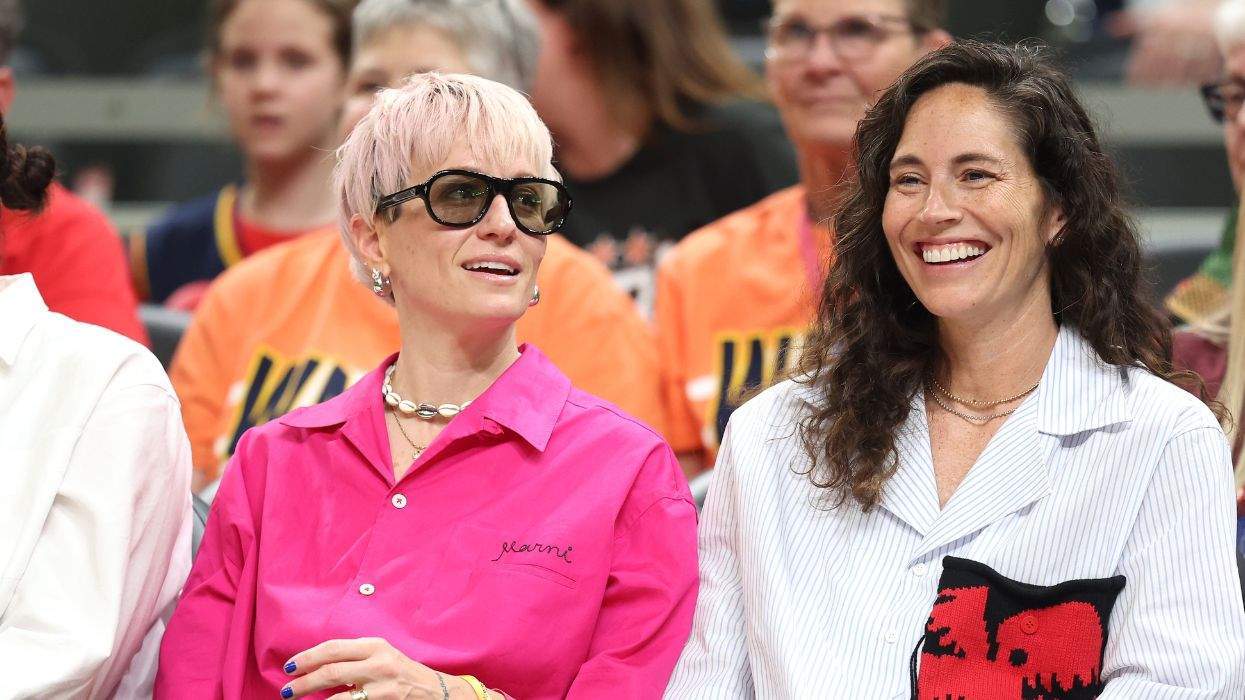 Megan Rapinoe and Sue Bird smiling and sitting at an event, one wearing sunglasses, others in the background.