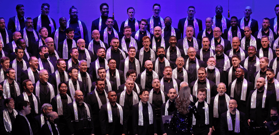 Members of the Gay Men\u2019s Chorus of Washington, DC perform together onstage wearing black outfits and silver scarves under purple stage lighting