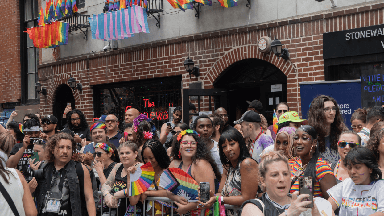 More than 35 people crowd the sidewalk outside the Stonewall Inn and Stonewall National Monument Visitor Center, on Christopher Street in Manhattan, watching NYC Pride March 2026. Dozens of small pride-themed flags hang on ironwork located below windows above the inn. This location in Greenwich Village is known as Stonewall.