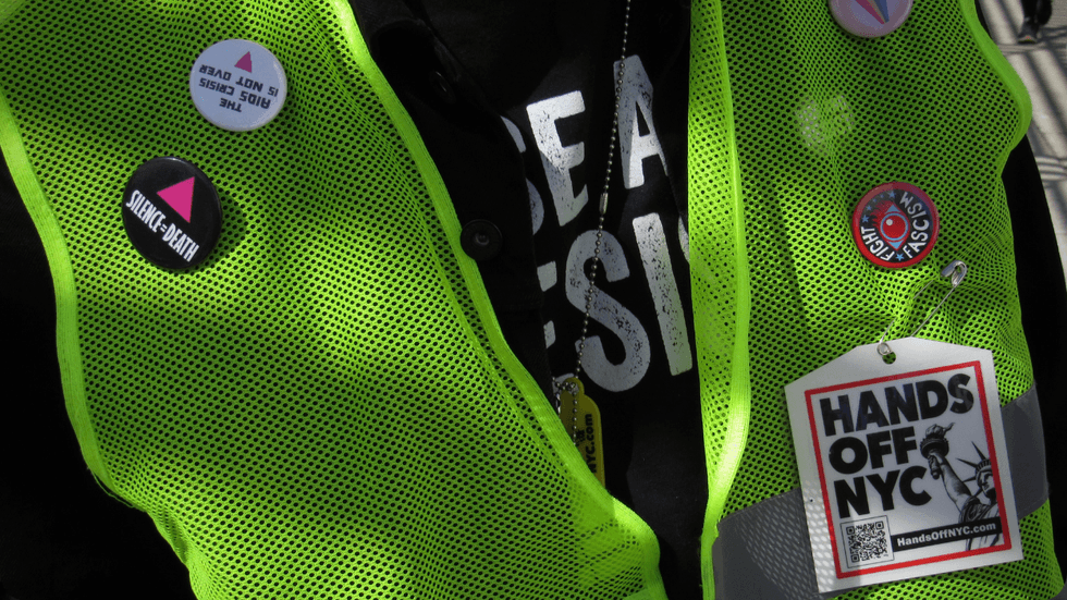 On a yellow, mesh vest, several activist pins and buttons are displayed. One reads "Silence = Death" with a pink triangle. Another reads "Hands Off NYC."