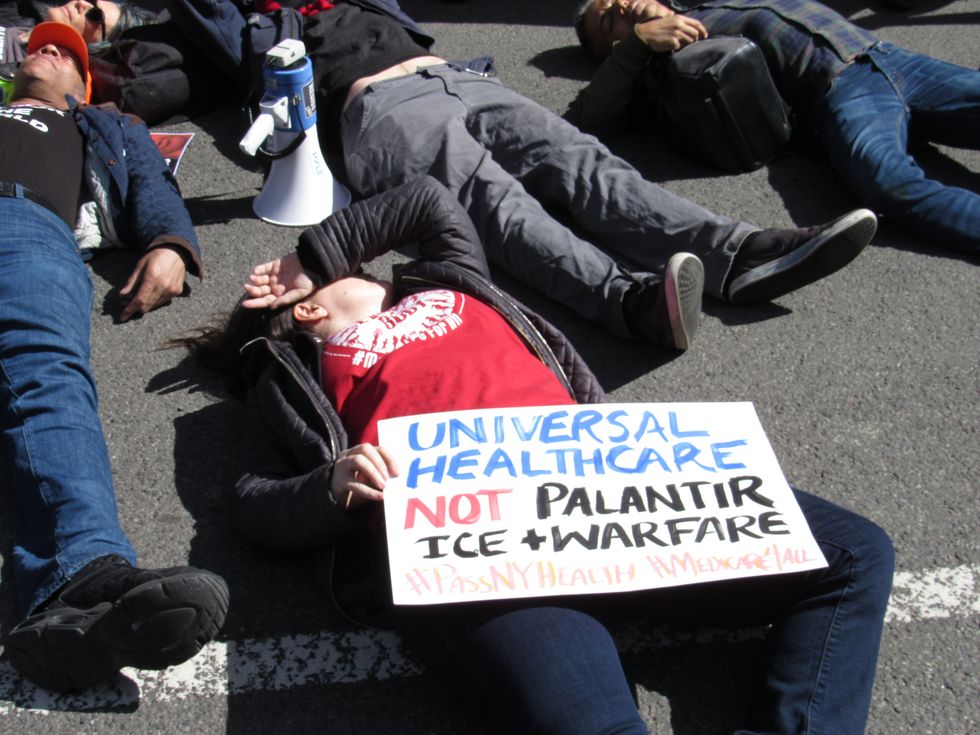 People lie in a city street. One at the center of the photo lies with a hand covering their face, holding a sign in their other hand that reads: "Universal health care, not Palantir, ICE & warfare."