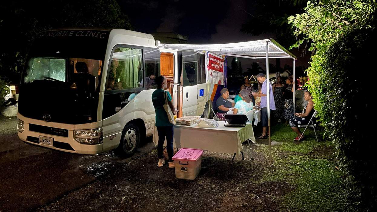 People sit under a roadside tent near a van and consult with members of the community.