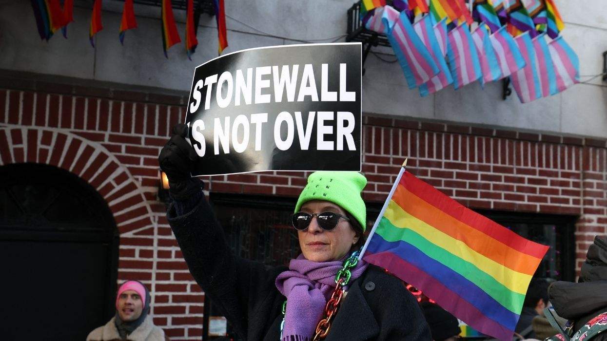 person standing in front of the stonewall inn holding pride flag and sign that reads stonewall is not over