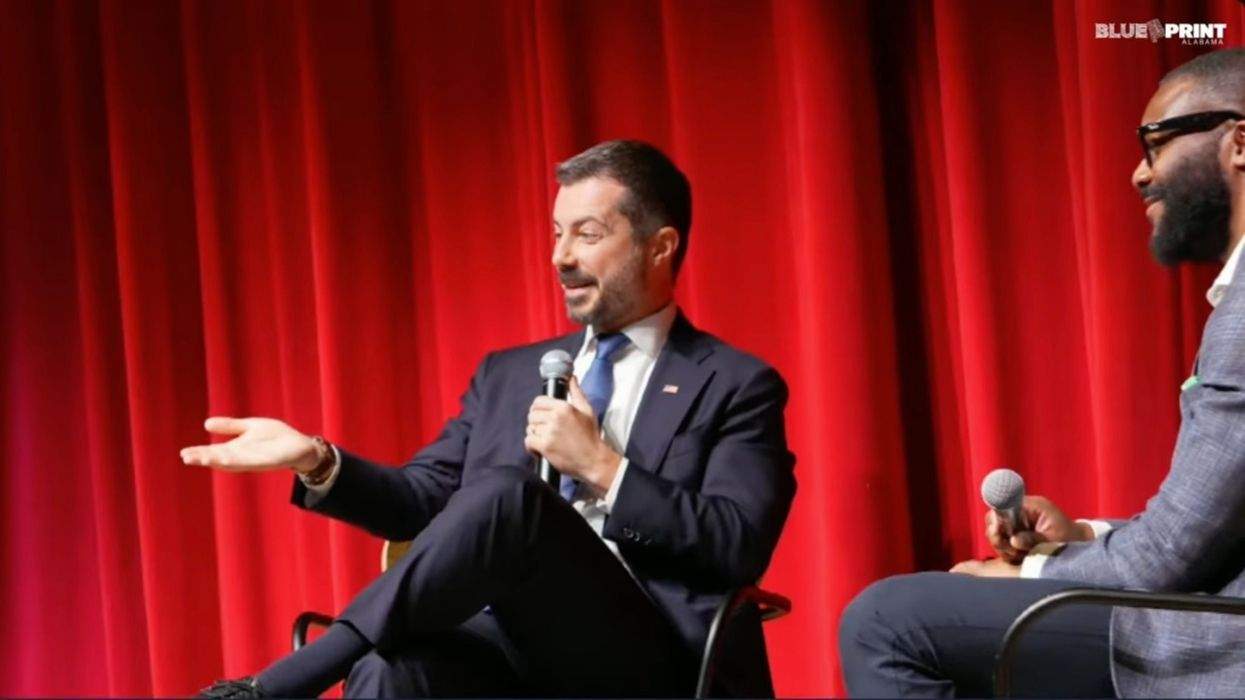 pete buttigieg onstage with a microphone in front of a red curtain alongside mayor randall woodfin of birmingham