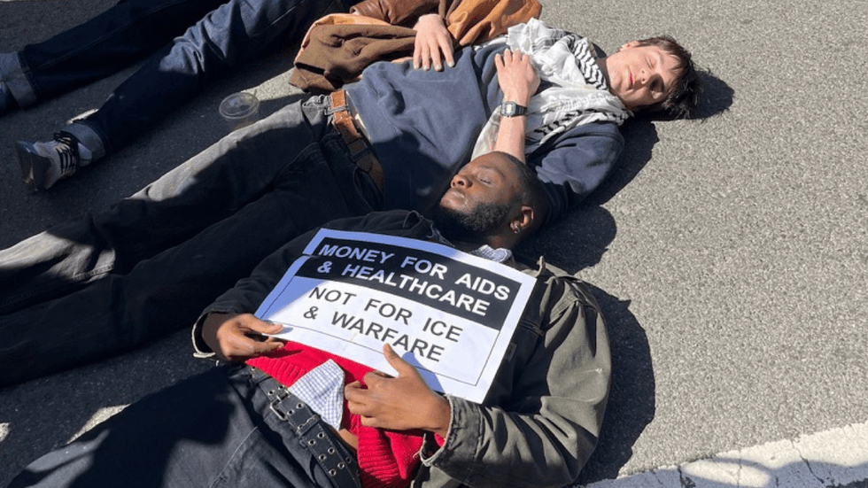 Protesters holding signs lie in a city street with their eyes closed.