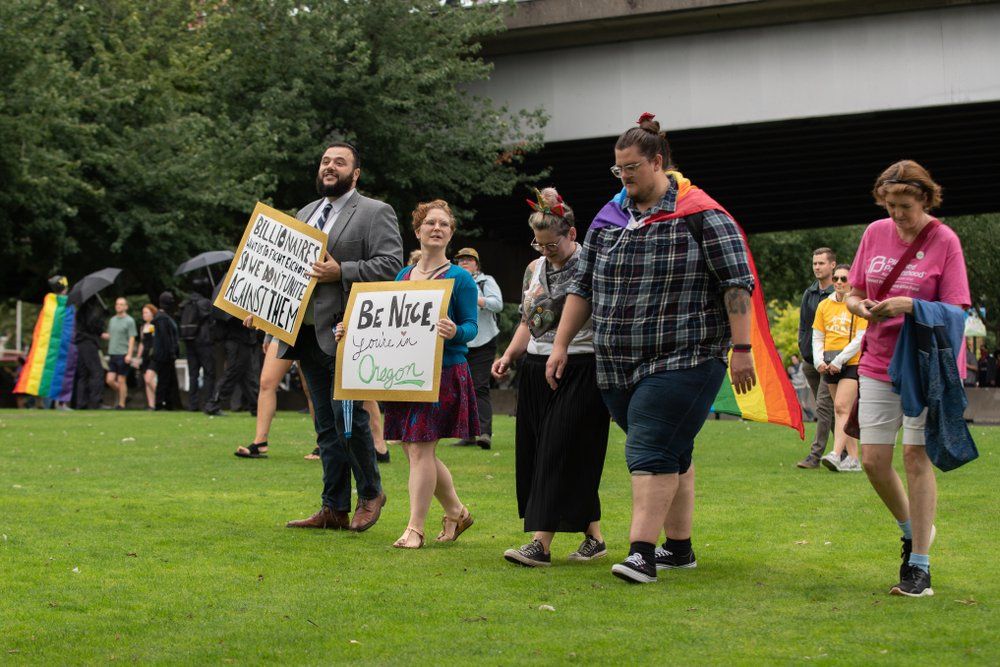 Protestors wear rainbow flag capes and carry a sign reading "Be nice, you're in Oregon"