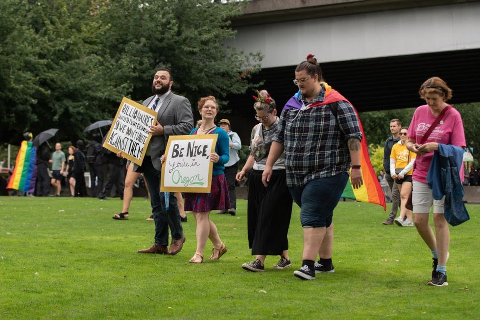 Protestors wear rainbow flag capes and carry a sign reading "Be nice, you're in Oregon"