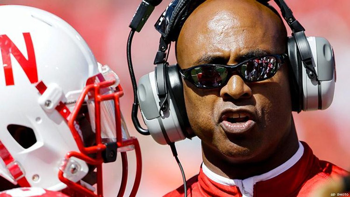 Ron Brown, football coach, wears sunglasses and headphones. A football player, whose face is obscured by a white helmet, stands to his right.