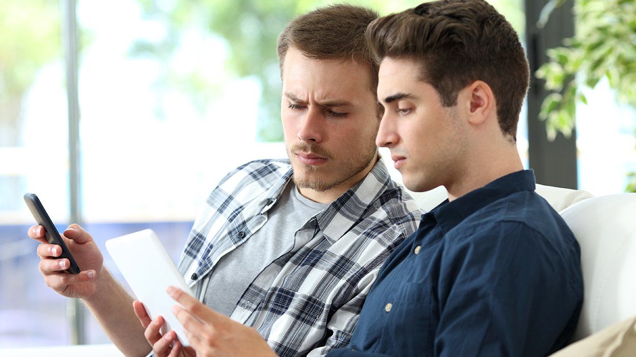 Serious gay men queer couple watching commercial tablet and phone sitting on a couch in the living room at home