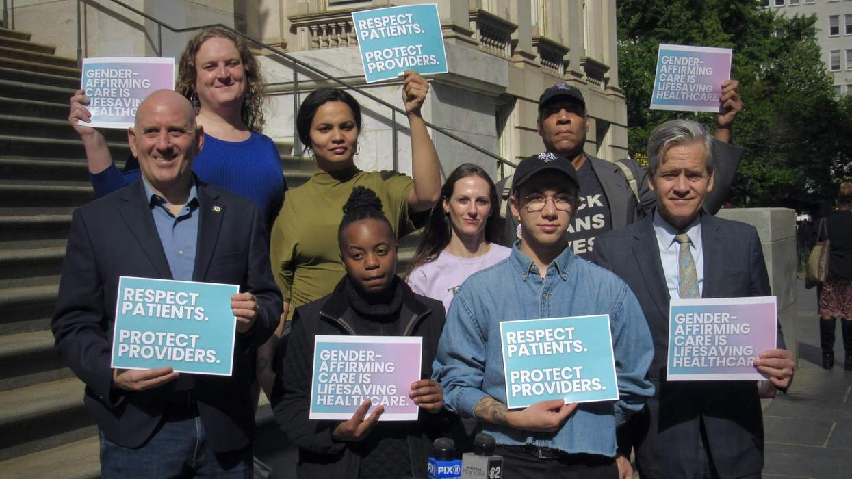 Several people stand along a staircase outside a tall government building. They stand around a pair of microphones holding signs that read "Respect Patients. Protect Providers." and "Gender-affirming care is lifesaving healthcare."