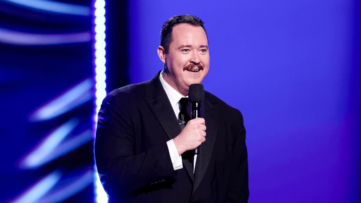 Shane Gillis wears a black suit and holds a microphone while speaking onstage against a blue and purple backdrop at the 2025 ESPY Awards.
