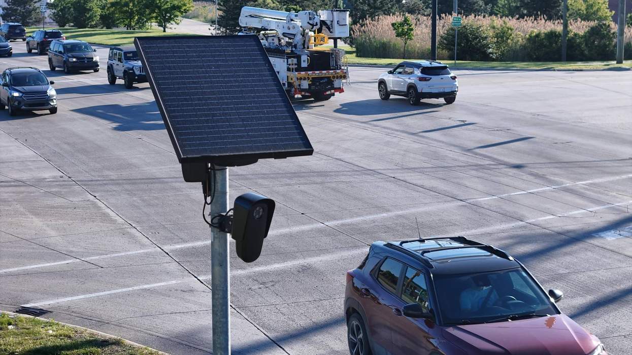Solar-powered camera at busy intersection with several cars passing by.