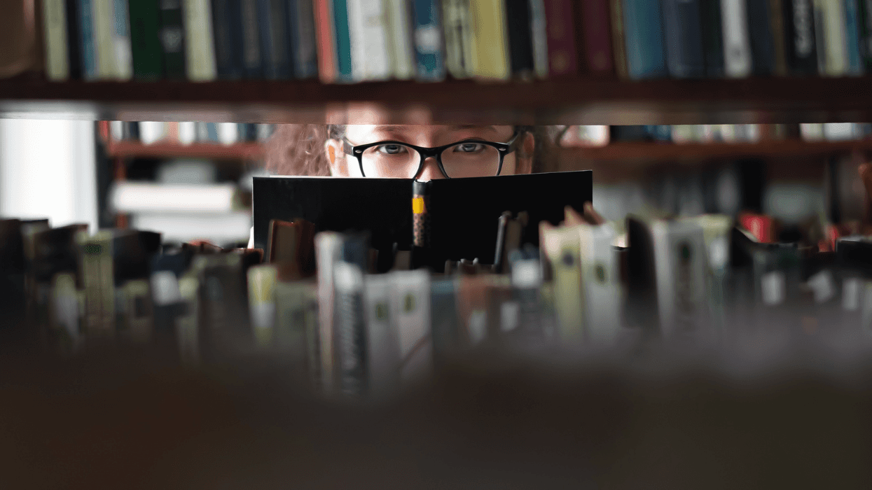 Student wearing glasses reads a book between shelves in a library