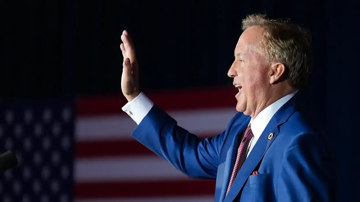 Texas Attorney General Ken Paxton, wearing a blue suit and red tie, raises his hand while speaking onstage in front of a blurred American flag backdrop