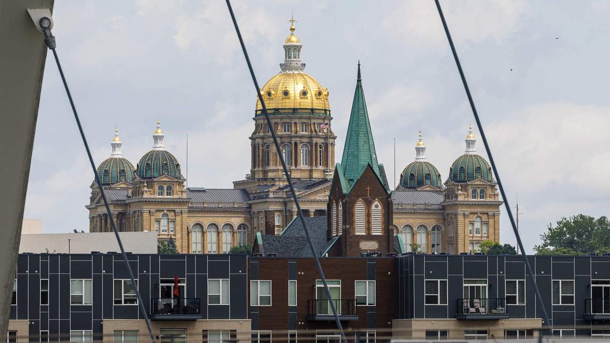 The golden dome of the Iowa State Capitol is visible through wire slats on a suspension bridge over a river. In front of the capitol sit a church and apartment building.