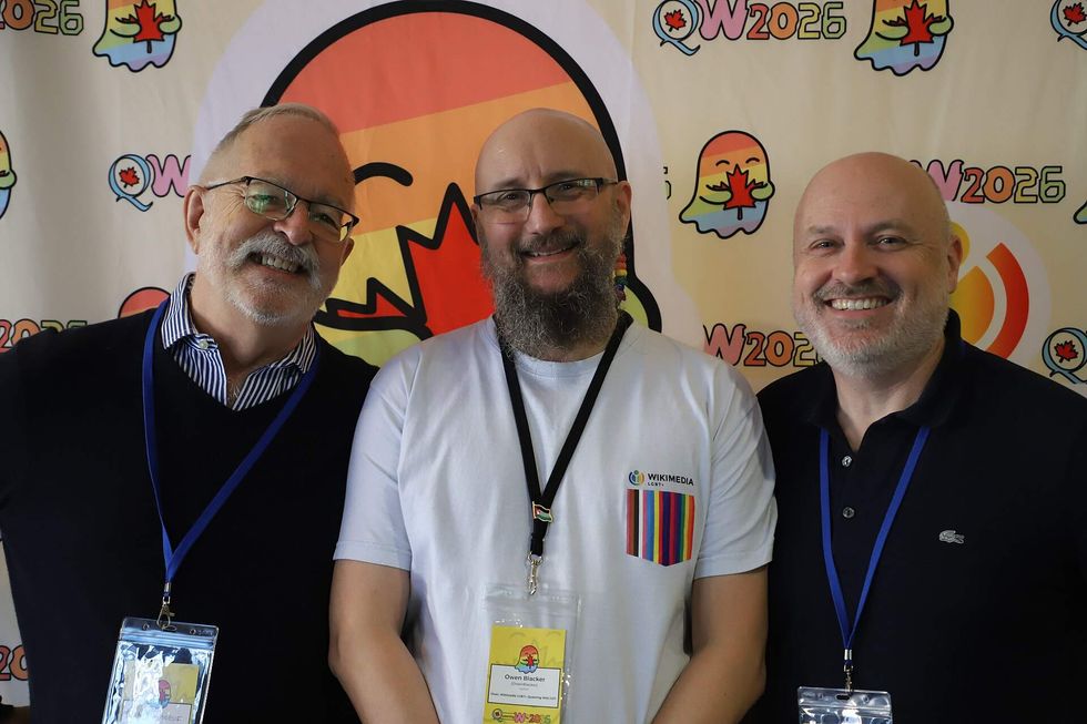 Three men stand in front of a colorful banner and smile toward the camera. They are each wearing conference lanyards.
