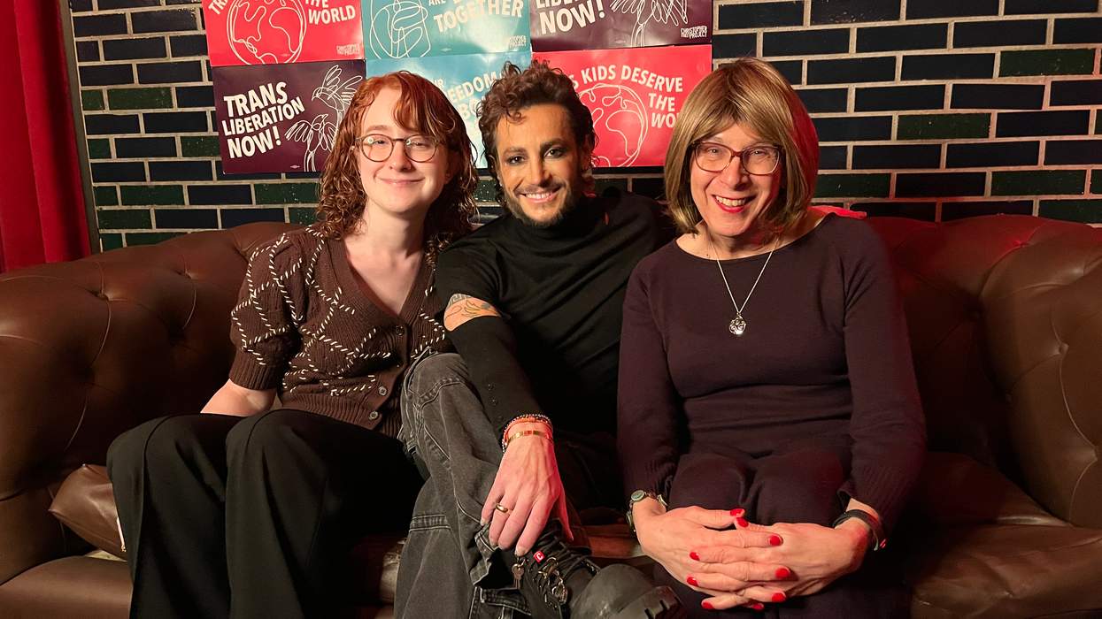 Three people sit on a brown couch wearing semi-formal attire. Behind them, posters read "Trans Liberation Now!" and "Our Freedoms Are Bound Together."