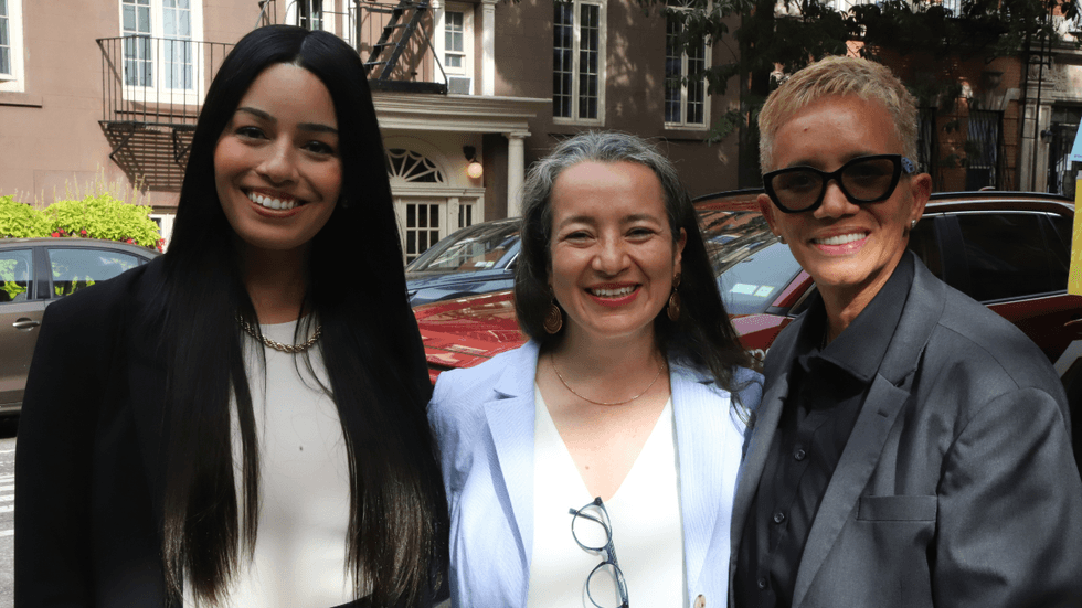Three women stand beside a city street and smile toward the camera.