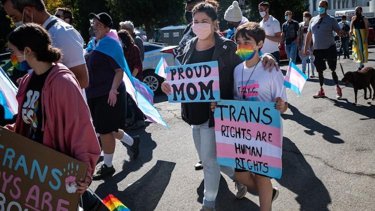 Trans youth march with woman holding "proud mom" sign in trans pride flag colors 