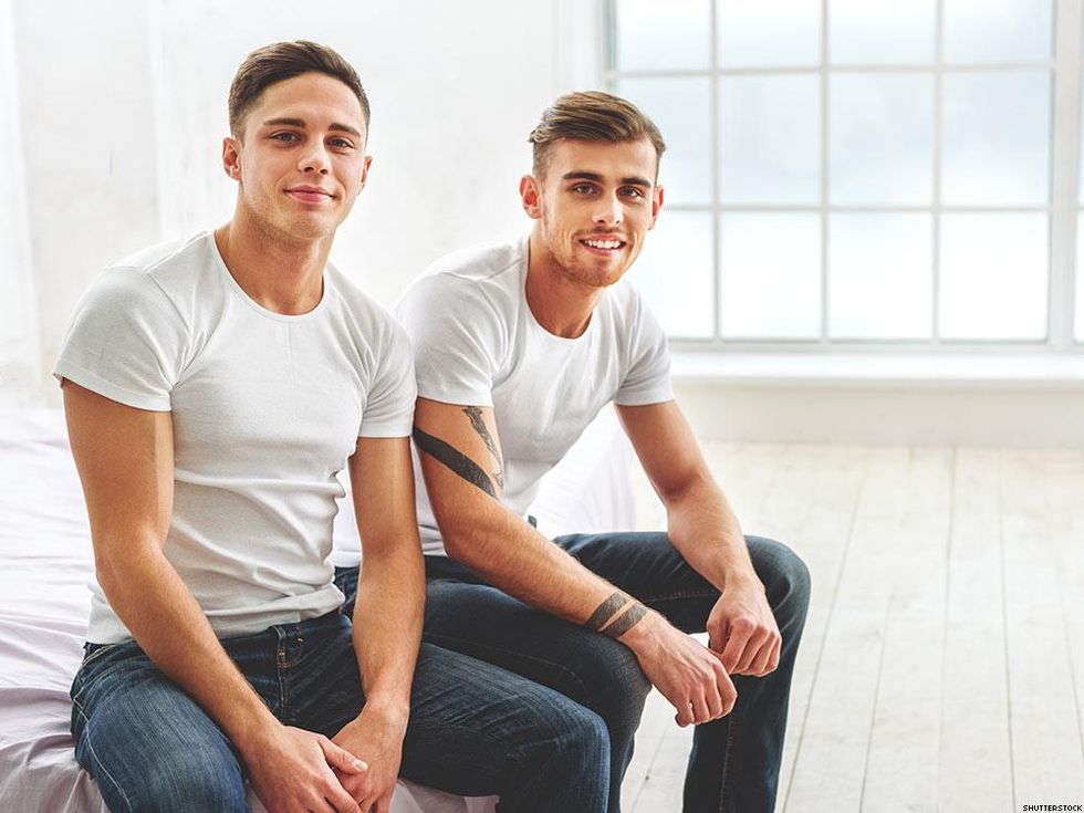 two guys in white tshirts and jeans sitting on the edge of a bed looking at the camera