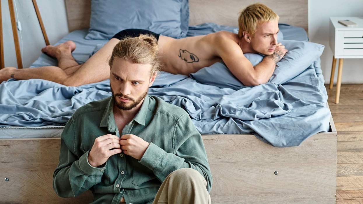 Two men in a bedroom; one sitting, distressed, the other lying on the bed, looking contemplative.