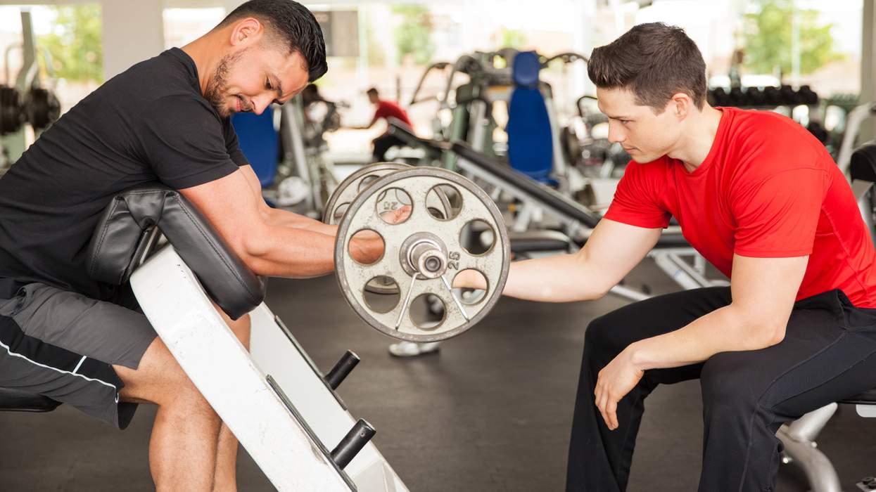 Two men in a gym, one assisting the other with a bicep curl.