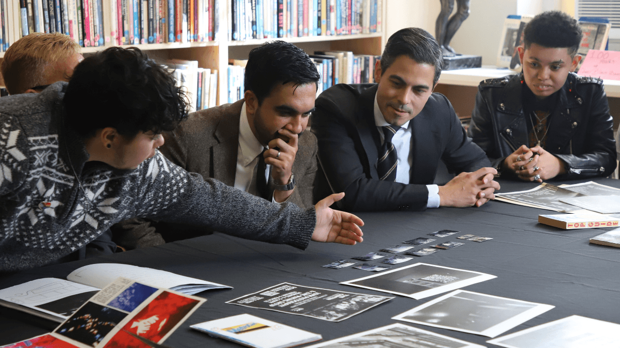Two men in suits and ties sit at a table with small pieces of artwork displayed. Teenagers sit around them and explain the artwork.
