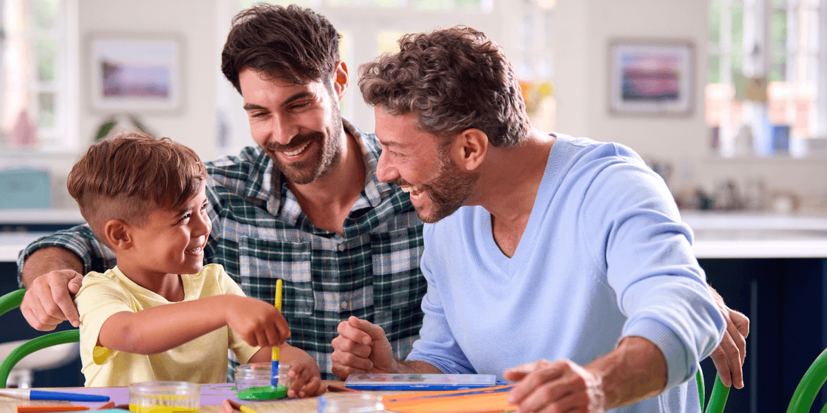 Two men smile and paint with a young boy at a table covered with colorful art supplies in a bright home.