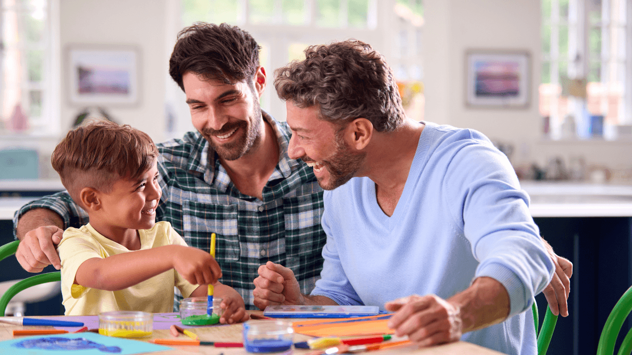 Two men smile and paint with a young boy at a table covered with colorful art supplies in a bright home.