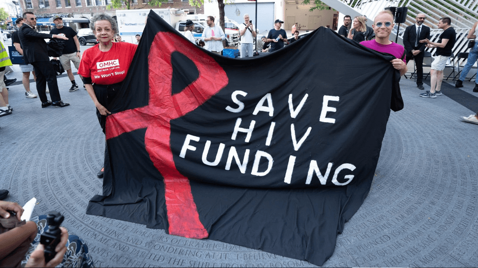 Two people hold up a large sign with a red ribbon that reads: "Save HIV funding."
