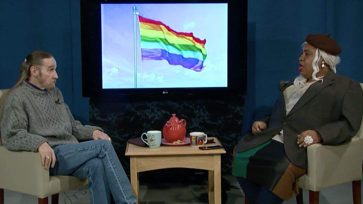 two people, one white and one black, sittting in chairs. the black person is dressed in drag. the whie person is dressed in jeans and a sweater