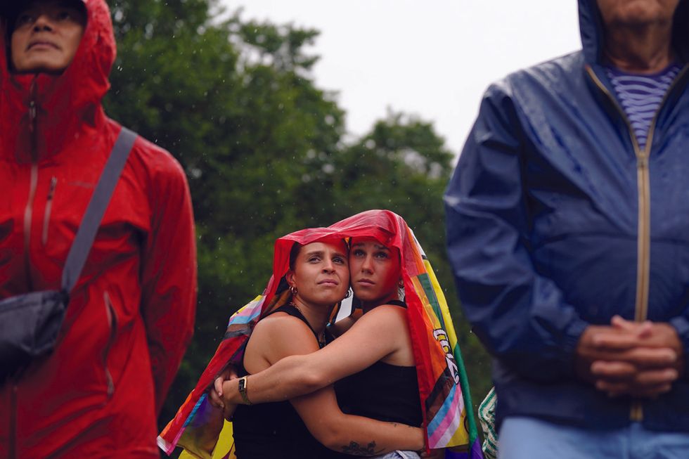 two women embrace with a rainbow colored flag draped over them