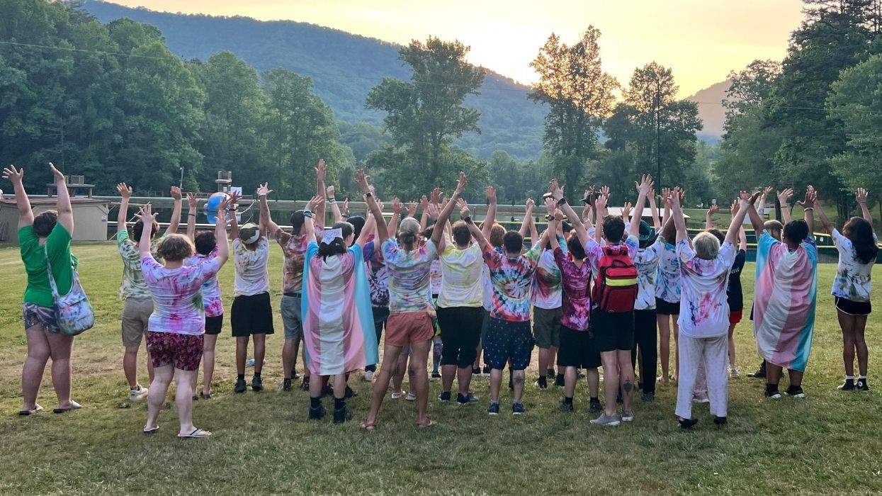 young people with arms raised in a group with trans flags and mountains and trees