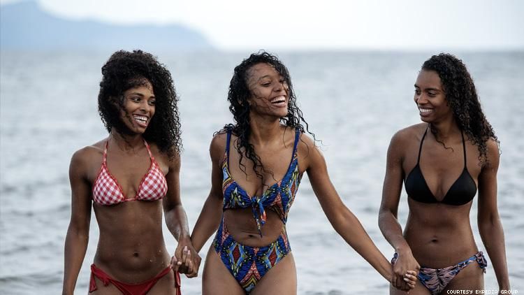 3 black women in swimsuits holding hands on a beach