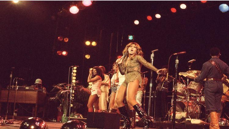 Tina Turner, a young fit Black woman in a dress, on stage in the 1970s